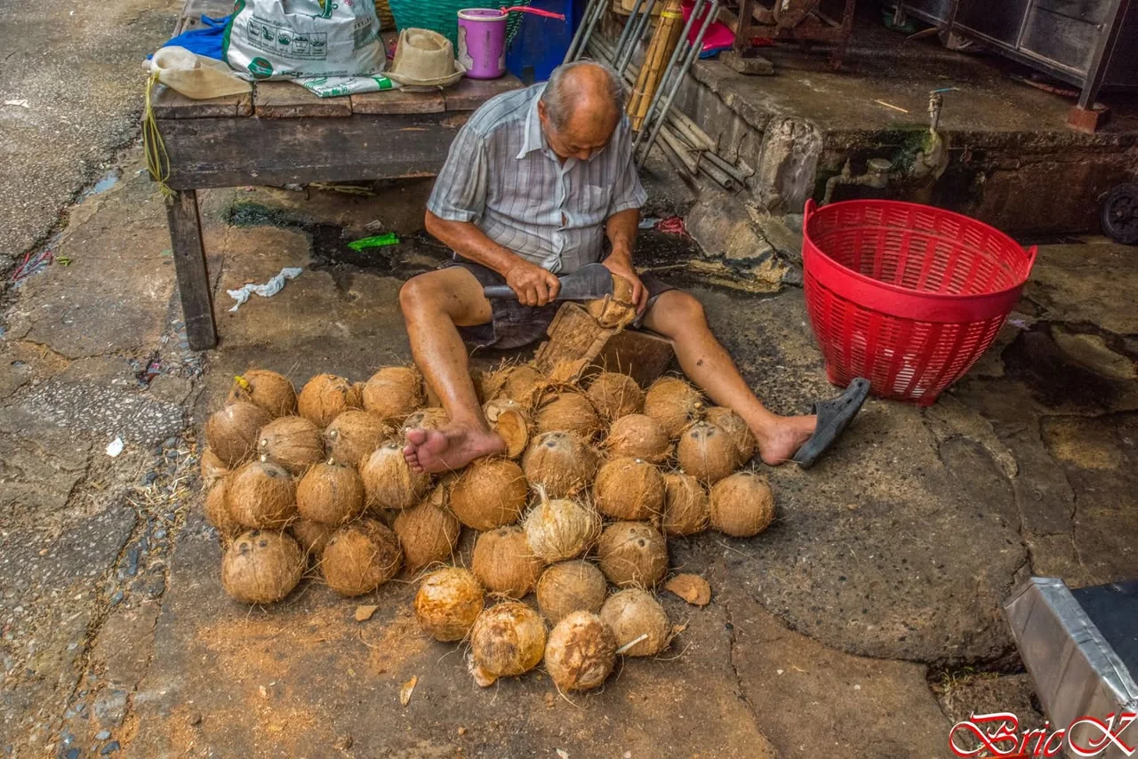 A worker prepares coconuts surrounded by tools, baskets and shells.