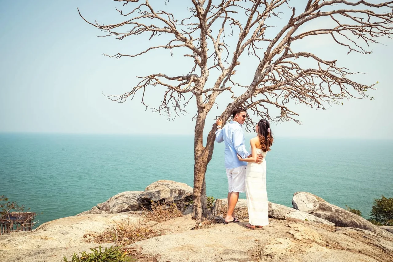 A couple stands beneath a coastal tree above calm blue water.