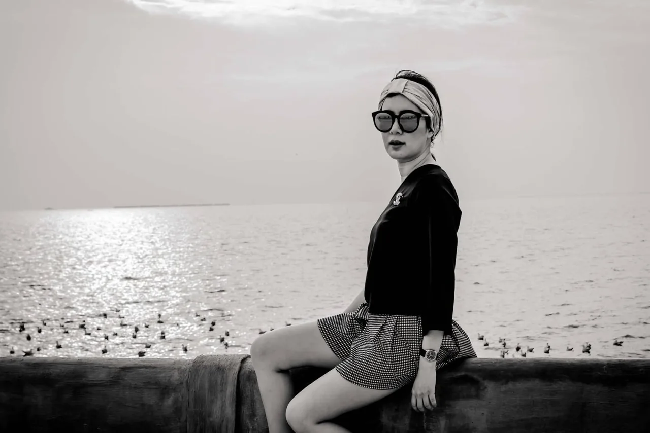 A monochrome portrait of a woman in sunglasses seated by the water.
