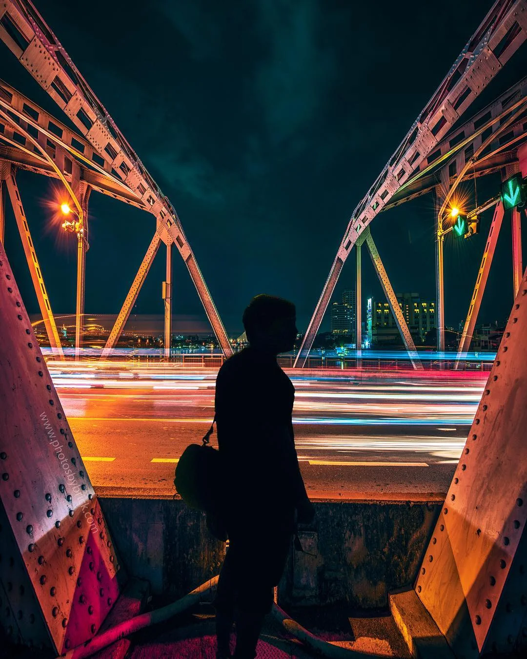 A silhouetted figure sits on a steel bridge beside orange and red light trails.