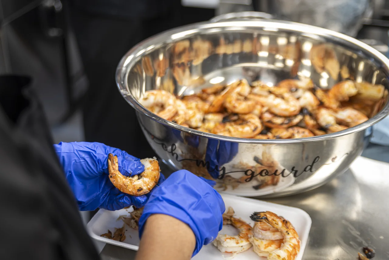 A gloved hand holds a prepared shrimp above a metal bowl in a kitchen.