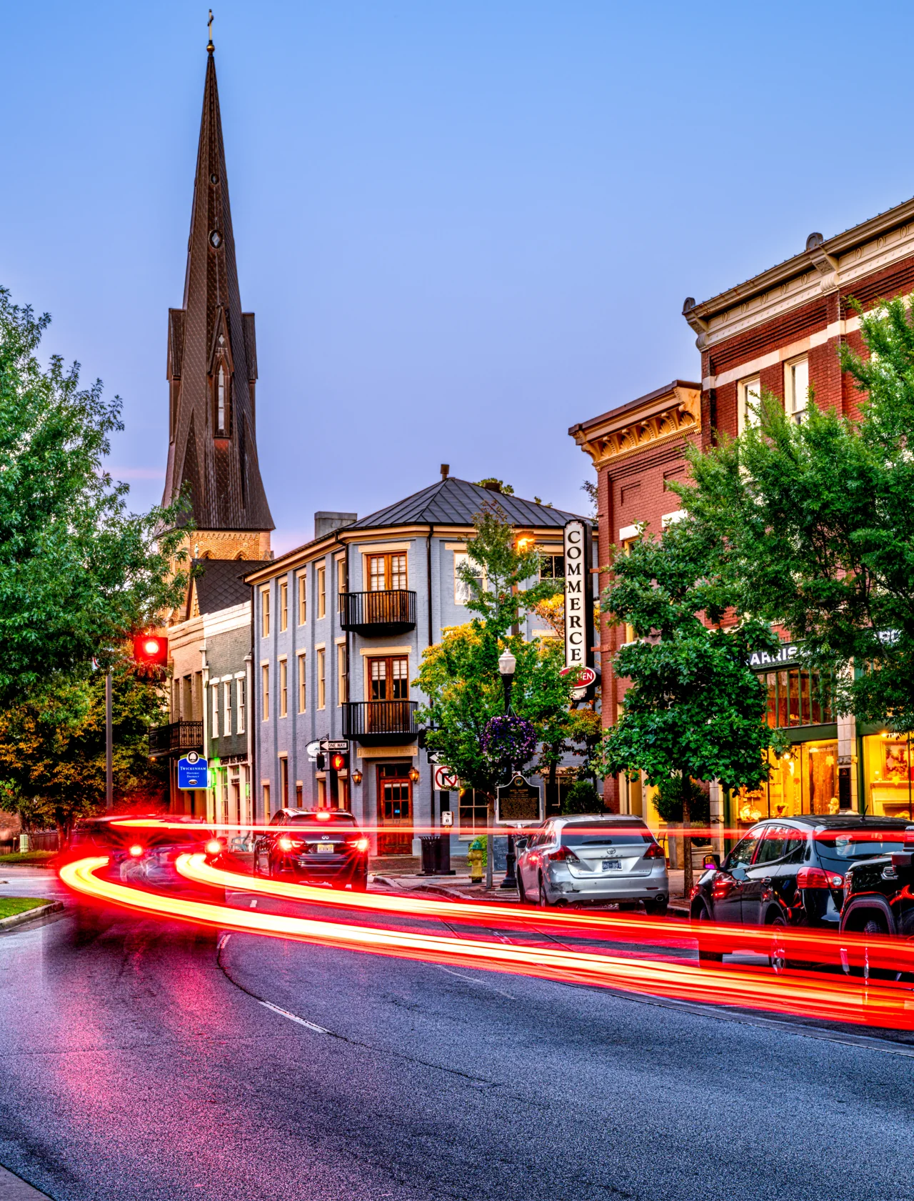 Red traffic trails lead through a city street beneath church spires at blue hour.
