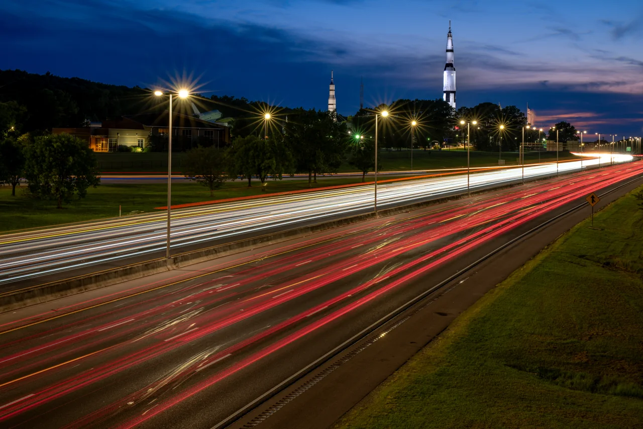 Highway light trails sweep toward a distant rocket-shaped landmark at dusk.