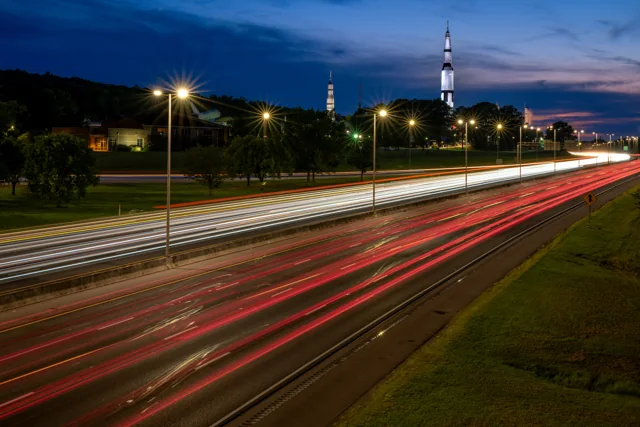 Highway light trails sweep toward a distant rocket-shaped landmark at dusk.