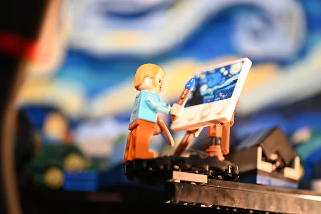 A small toy painter works at an easel in warm light against a blue painting.