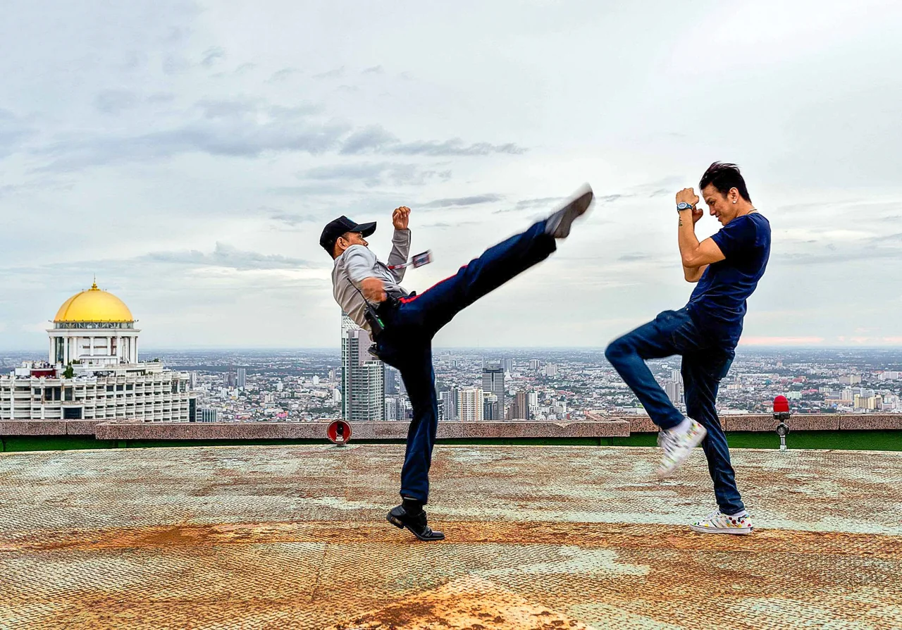 A martial artist makes a high kick toward another performer on a rooftop.