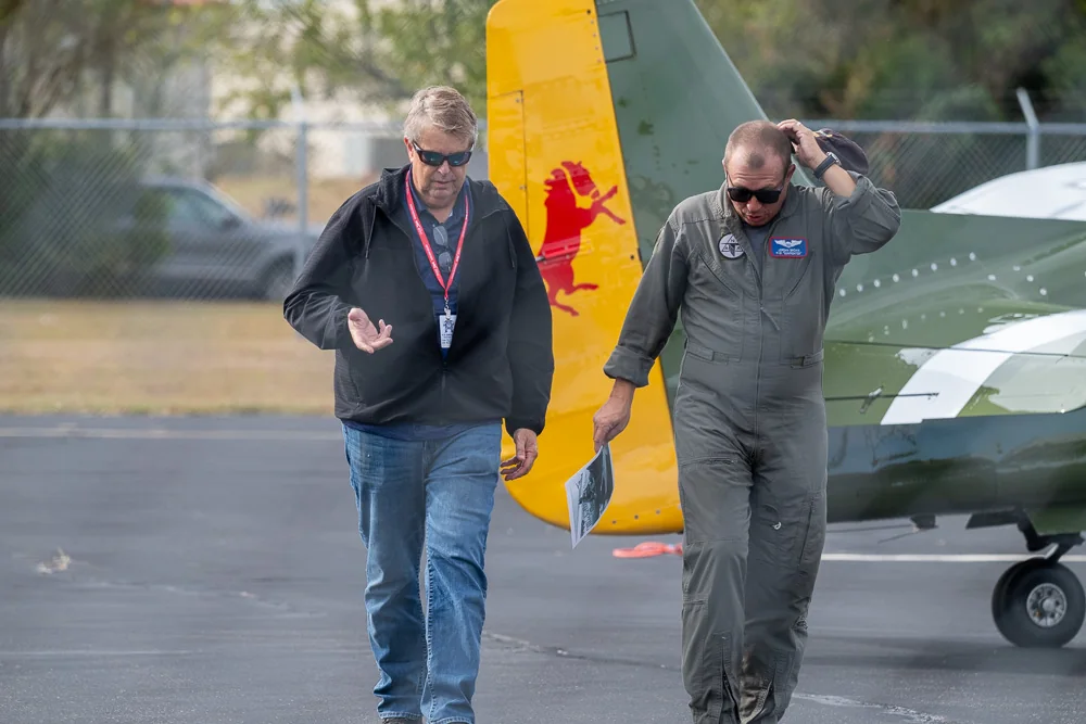 Two people walk beside the yellow tail of an aircraft.